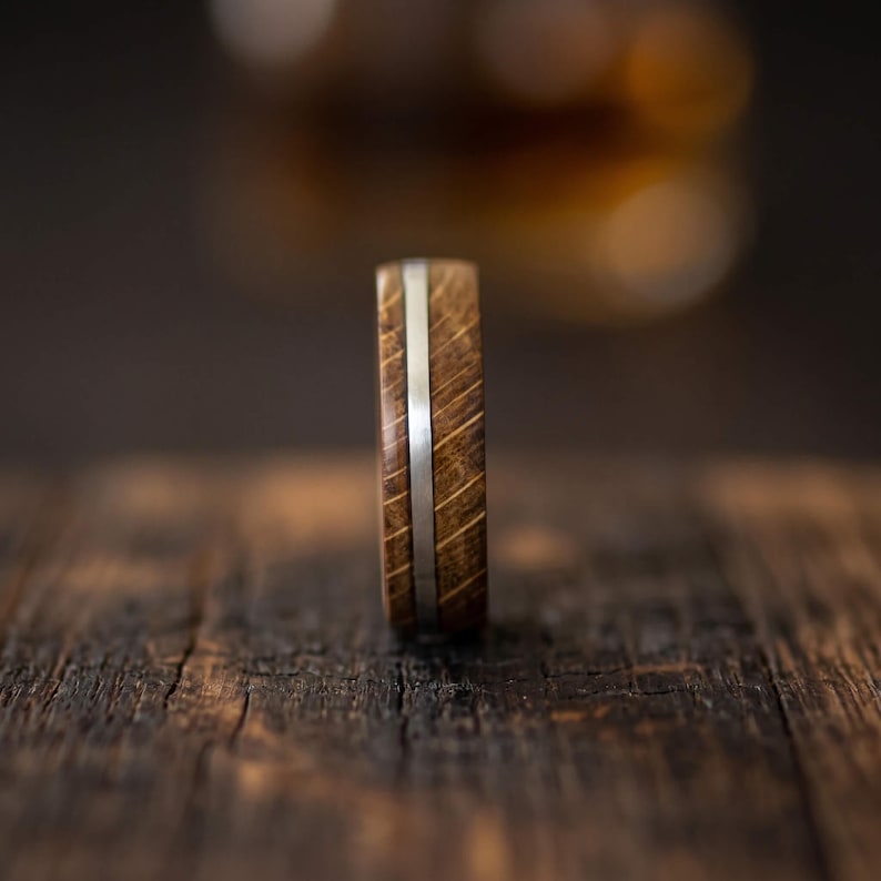 A ring crafted from brown oak wood featuring a smooth, polished silver inlay that runs horizontally around the center of the band. The wood grain is visible, showing a natural texture and deep brown tones throughout the ring.
