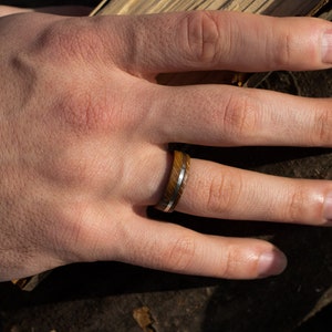 A ring crafted from brown oak wood featuring a smooth, polished silver inlay that runs horizontally around the center of the band. The wood grain is visible, showing a natural texture and deep brown tones throughout the ring.