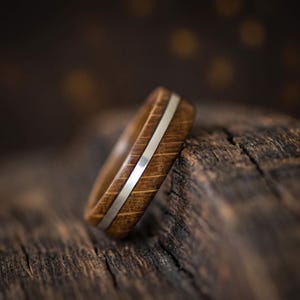 A ring crafted from brown oak wood featuring a smooth, polished silver inlay that runs horizontally around the center of the band. The wood grain is visible, showing a natural texture and deep brown tones throughout the ring.