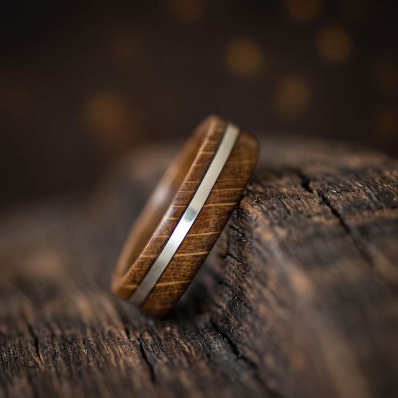 A ring crafted from brown oak wood featuring a smooth, polished silver inlay that runs horizontally around the center of the band. The wood grain is visible, showing a natural texture and deep brown tones throughout the ring.