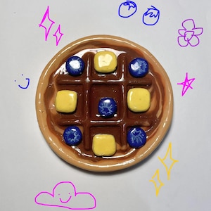 May include: A ceramic waffle with a brown grid pattern, topped with yellow squares and blue circles. The waffle is surrounded by a light brown border. The background has doodles of stars, flowers, and a cloud.