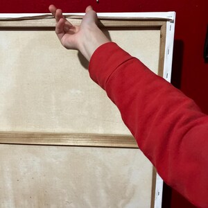 May include: A large, rectangular canvas art piece with a white border and wooden frame. The canvas is a light beige colour. A person's red sleeve and hand are reaching towards the canvas, suggesting it's being hung on a red wall.