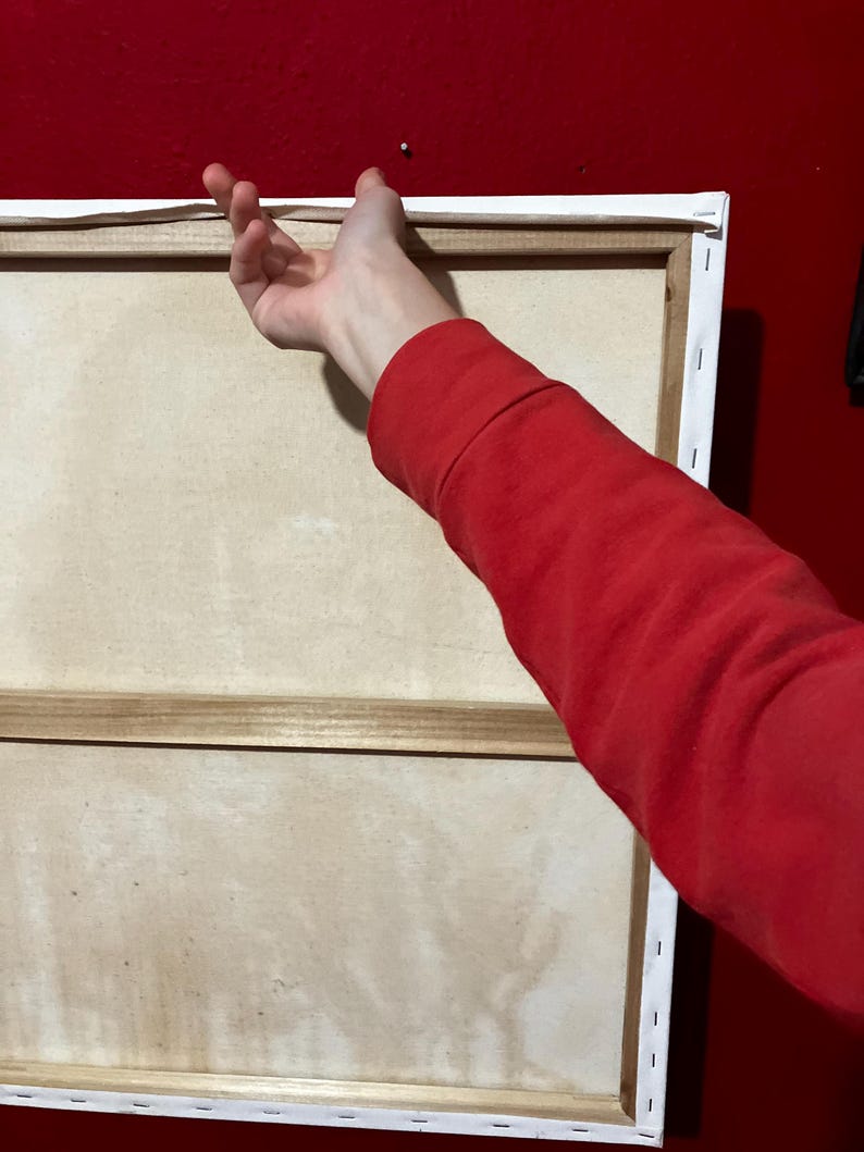 May include: A large, rectangular canvas art piece with a white border and wooden frame. The canvas is a light beige colour. A person's red sleeve and hand are reaching towards the canvas, suggesting it's being hung on a red wall.