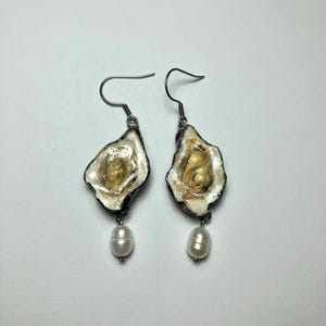 May include: Handmade earrings with oyster shell designs and a pearl drop. The earrings have silver-tone metal hooks and a white and beige colour scheme. The oyster shells have a textured appearance.