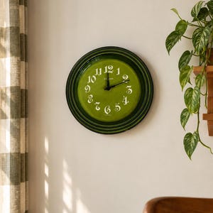 May include: A round wall clock with a glossy olive green face and a dark green rim. The clock features white stylized numerals from one to twelve and two black hands. The center of the clock face displays the text Le Creuset in white lettering.