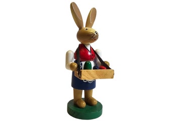May include: A wooden Easter bunny figurine with brown fur, a white shirt, a red vest, and a blue skirt. The bunny is holding a tray with colorful Easter eggs.