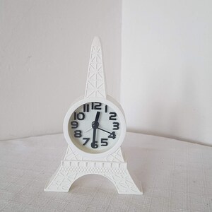 Puede incluir: Reloj despertador blanco con forma de Torre Eiffel con agujas y números negros.