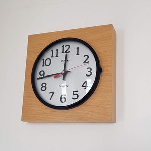 May include: A simple, round, black-framed clock with white face and black numbers. The clock is mounted on a light wood square frame.