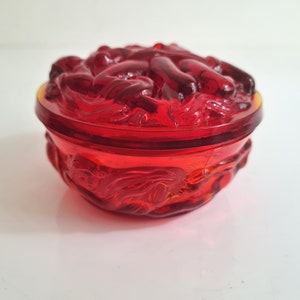 May include: A red glass trinket box with a lid featuring a raised design of intertwined figures. The box is round and has a smooth, polished finish.