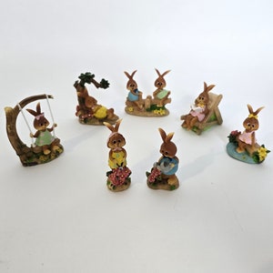 Op de afbeelding: Een set van zeven bruine konijnenfiguren, elk met verschillende poses en accessoires. De konijnen zitten op schommels, banken en boomstammen, en sommigen houden bloemen of gieters vast. De figuren zijn gemaakt van hars en zijn geschilderd in felle kleuren.