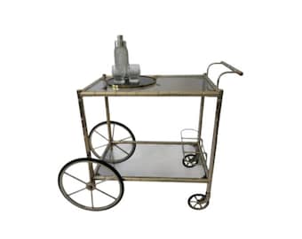 May include: A two-tiered silver bar cart with glass shelves and wheels. The cart has a silver bottle opener and a glass tray on the top shelf.