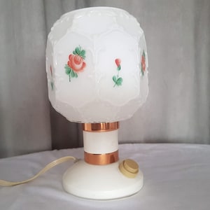 May include: White table lamp with a frosted glass shade featuring a floral design of red roses and green leaves. The lamp has a copper base with a white switch.