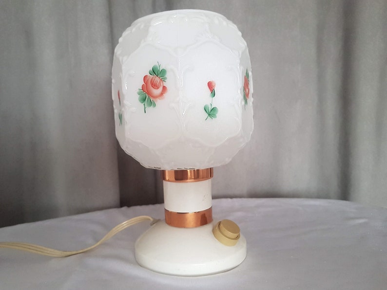 May include: White table lamp with a frosted glass shade featuring a floral design of red roses and green leaves. The lamp has a copper base with a white switch.