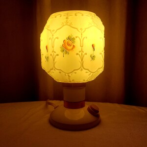 May include: A white table lamp with a frosted glass shade featuring a floral design. The shade has a raised pattern and is illuminated from within. The lamp has a white base with a brown accent band and a cord with an on/off switch.