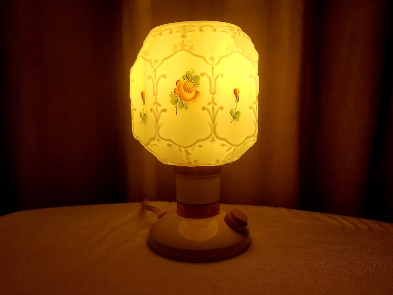 May include: A white table lamp with a frosted glass shade featuring a floral design. The shade has a raised pattern and is illuminated from within. The lamp has a white base with a brown accent band and a cord with an on/off switch.