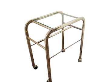 May include: A gold metal rolling cart with a glass top and a lower shelf. The cart has four wheels and is made of a sleek, modern design.