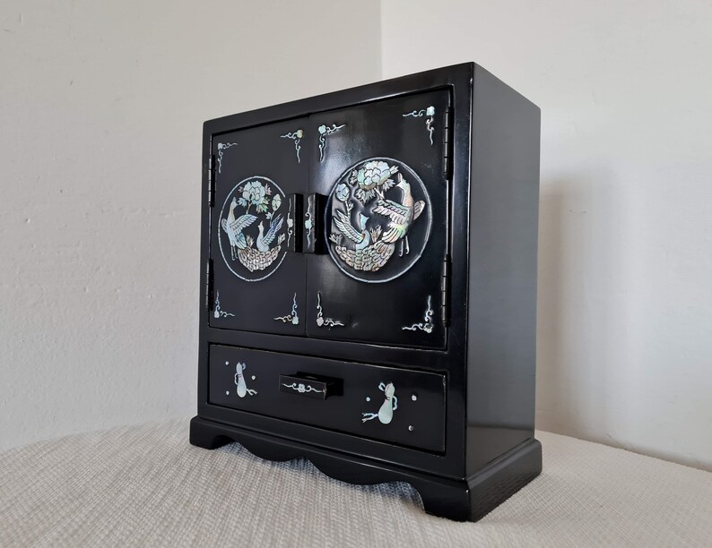 May include: A black wooden jewelry box with a single drawer and two doors. The doors are decorated with a circular design of birds and flowers made from mother of pearl. The drawer has a mother of pearl design of a cat on each side.