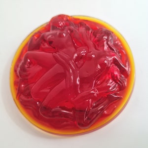 May include: A red glass paperweight with a design of three figures intertwined. The figures are in a circular shape and the paperweight has a yellow rim.