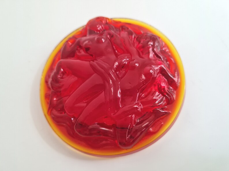 May include: A red glass paperweight with a design of three figures intertwined. The figures are in a circular shape and the paperweight has a yellow rim.