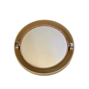 May include: A round mirror with a light brown wooden frame. The mirror has a circular reflective surface and is held in place by two silver metal brackets. The frame has a smooth, matte finish.