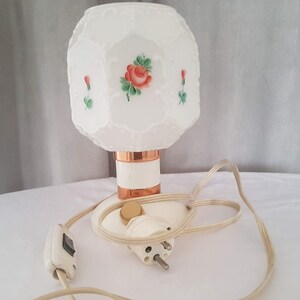 May include: White table lamp with a white and copper base and a white octagonal shade with a floral design. The shade has a raised pattern and is decorated with red roses and green leaves. The lamp has a white cord and a white switch.