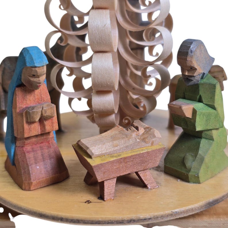 May include: A wooden nativity scene with a carved wooden tree, a manger, and three figures. The figures are painted in shades of blue, red, green, and brown. The manger is made of wood and has a yellow trim.