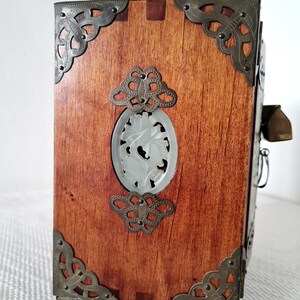 May include: A wooden box with ornate metal corners and a carved jade panel on the front. The box has a dark brown stain and is decorated with intricate metalwork.