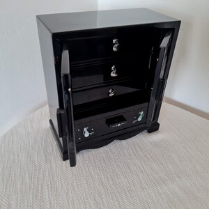 May include: Black wooden jewelry box with four drawers and two doors. The doors are open and the drawers are decorated with small white flowers.