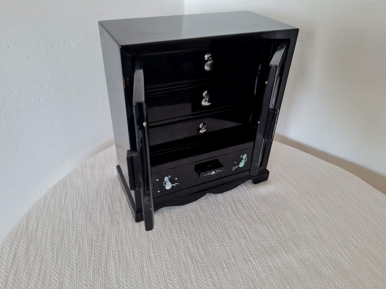 May include: Black wooden jewelry box with four drawers and two doors. The doors are open and the drawers are decorated with small white flowers.