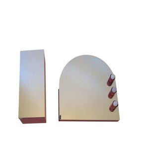 May include: A modern vanity mirror set with a rectangular mirror and an arched mirror. The mirrors have a light-colored reflective surface and are framed with a reddish-brown border. The arched mirror has three cylindrical, reddish-brown knobs.