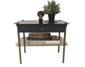 May include: A black side table with a glass top and a drawer. The table has a lower shelf and gold metal legs. A book titled "De Natuur" is on the shelf. A small figurine and a potted plant are on the top of the table.
