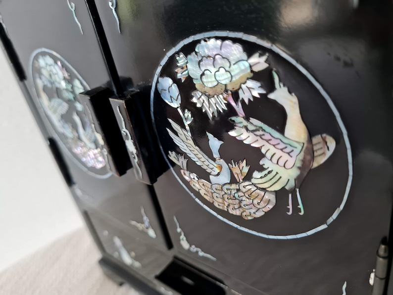 May include: Black wooden cabinet with intricate mother-of-pearl inlay depicting a pair of birds and a flower. The cabinet has two doors and multiple drawers.