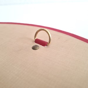 May include: A close-up of a red leather-bound journal with a gold metal ring attached to the top. The journal has a beige fabric cover.