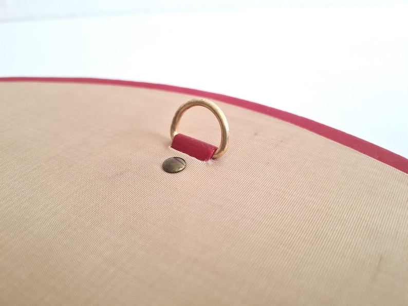 May include: A close-up of a red leather-bound journal with a gold metal ring attached to the top. The journal has a beige fabric cover.