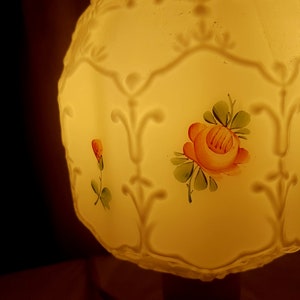 May include: A white glass lampshade with a floral design. The lampshade has a yellow glow and features a pink rose with green leaves.