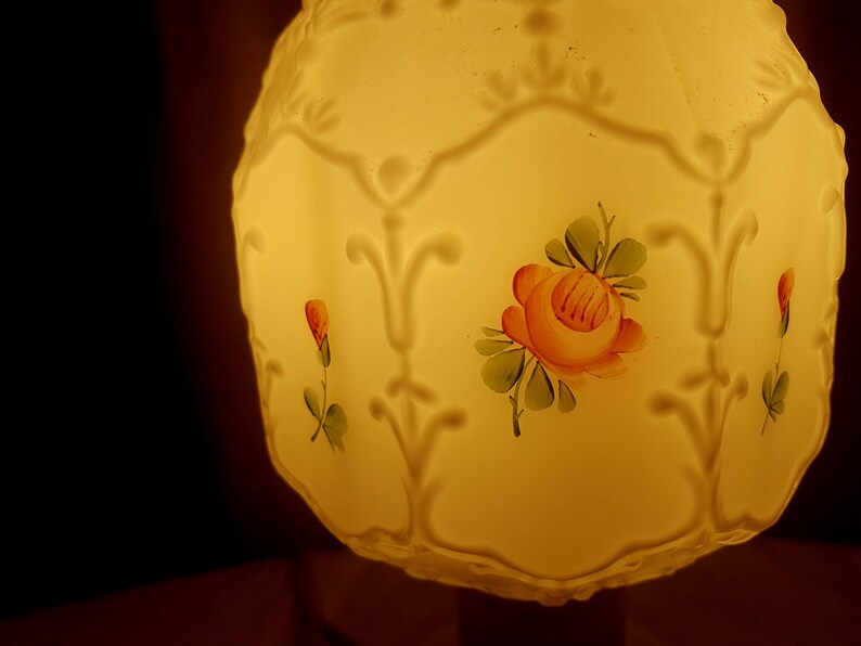 May include: A white glass lampshade with a floral design. The lampshade has a yellow glow and features a pink rose with green leaves.