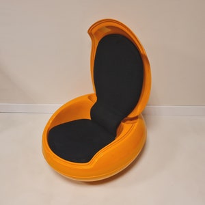 May include: A bright orange egg-shaped chair with a black fabric seat and a curved back. The chair is open and ready to be sat in.