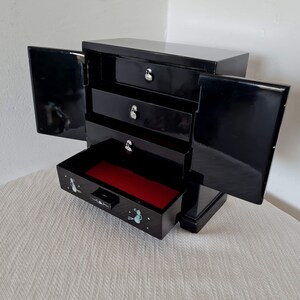 May include: Black wooden jewelry box with three drawers and a hinged lid. The box has a red velvet interior and a small drawer with a floral design.