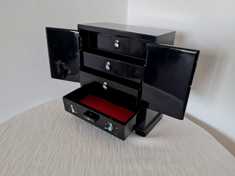 May include: Black wooden jewelry box with three drawers and a hinged lid. The box has a red velvet interior and a small drawer with a floral design.