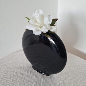 May include: A glossy black vase with a unique, asymmetrical shape. The vase is filled with white roses and green leaves. The vase has a textured lower half and a smooth, reflective upper half. A decorative floral arrangement.