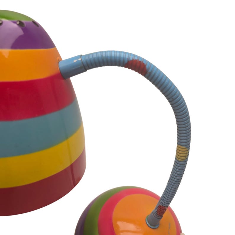 May include: A colorful desk lamp with a rainbow striped shade and a flexible blue neck. The base is also multi-colored, featuring a vibrant mix of red, orange, yellow, green, and purple. The lamp is designed for desk use.