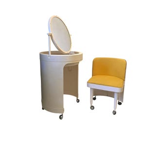 Può includere: Un set da toeletta vintage. La toeletta cilindrica color crema ha uno specchio rotondo. La sedia abbinata ha una seduta gialla. Entrambi i pezzi hanno le ruote.