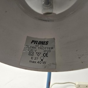 May include: A close-up of a white and blue desk lamp. The lamp shade has a label that reads "PYLONES GLOBE TROTTER 230 V IP20 E27 max 40 W". The lamp has a blue, flexible neck and a white power cord.