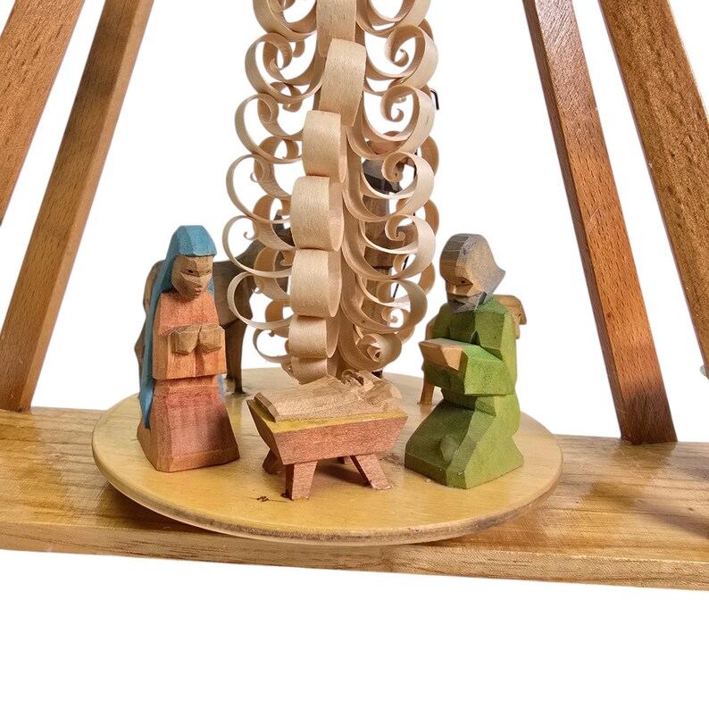 May include: A wooden nativity scene with a carved wooden tree, a manger with a baby Jesus, and two figures kneeling in prayer. The figures are painted in bright colors, including red, blue, green, and yellow.