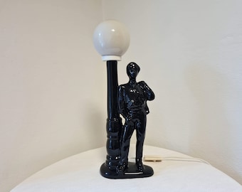 May include: Black ceramic table lamp with a white globe shade. The lamp features a figurine of a person standing next to a lamp post.