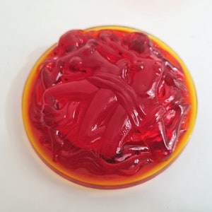 May include: A red glass paperweight with a swirling design. The paperweight is set in a clear glass base with a yellow rim.