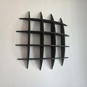 Op de afbeelding: Een donkerbruin wandrek in rasterstijl met meerdere compartimenten. Het rek is gemaakt van hout en heeft een geometrisch ontwerp. Het rek is aan een witte muur bevestigd en toont zijn unieke vorm en functionaliteit.