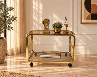 Puede incluir: Un carrito de metal dorado con dos estantes rectangulares de cristal. El estante superior sostiene un reloj redondo dorado, una pequeña planta verde en maceta y una figura de pájaro dorada. El estante inferior contiene el libro The Kinfolk Home.