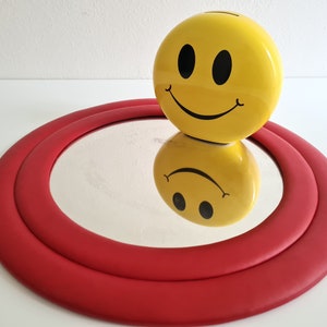 May include: A yellow smiley face piggy bank sits on top of a red circular mirror with a double rim.
