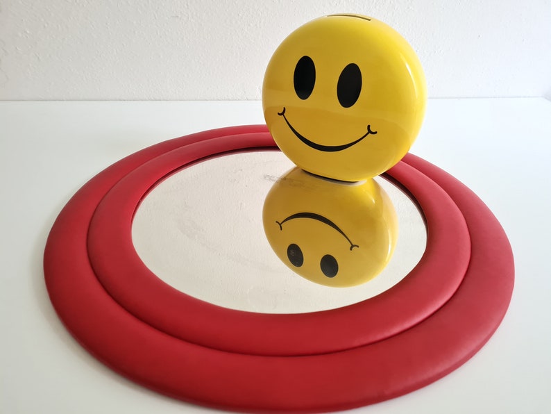 May include: A yellow smiley face piggy bank sits on top of a red circular mirror with a double rim.